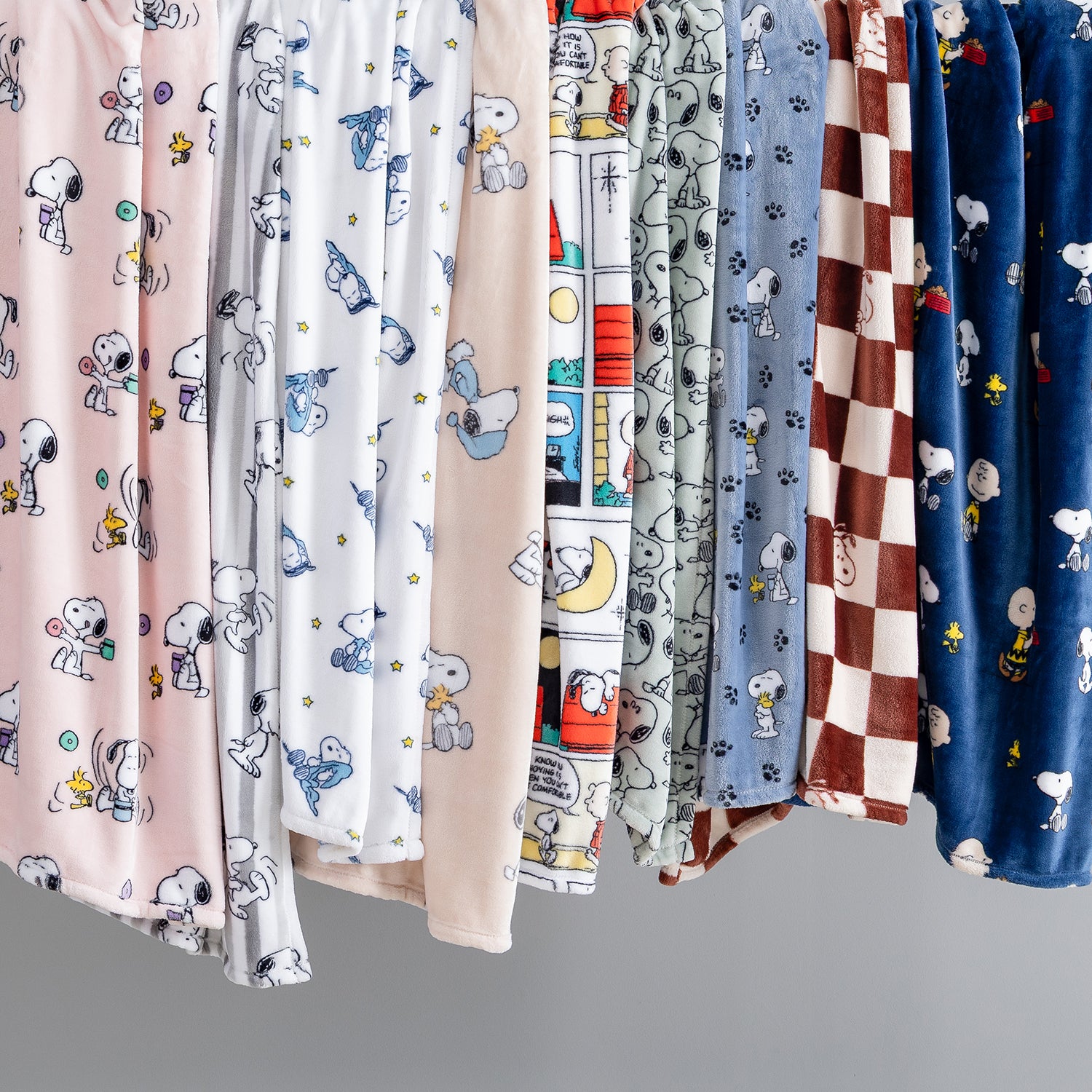 A row of plush throw blankets hanging side-by-side, featuring various cartoon prints including Snoopy and Woodstock on pink, white, beige, gray, blue, and brown patterns.