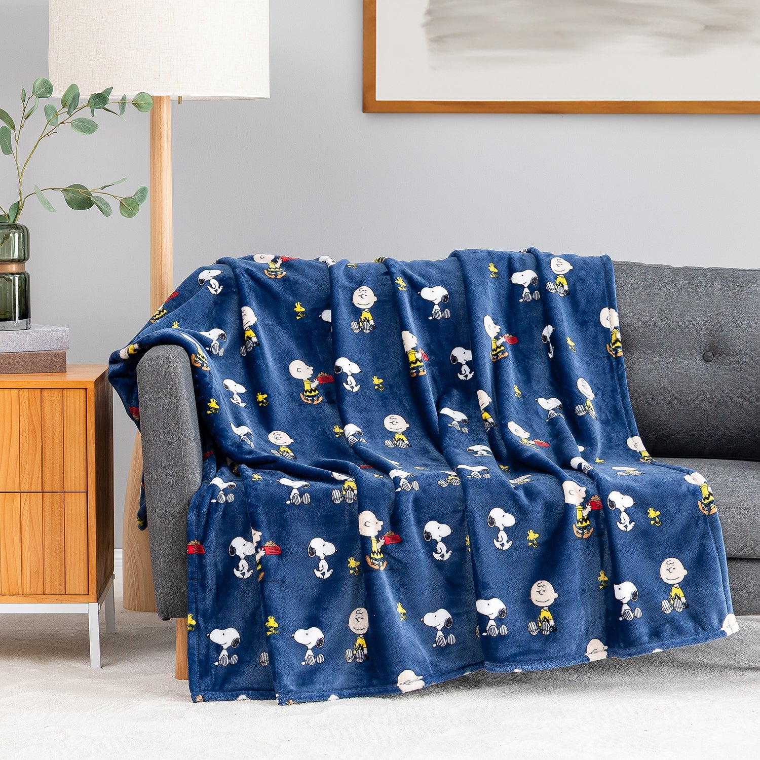 A navy blue fleece throw blanket featuring a repeating cartoon print of Snoopy, Woodstock, and Charlie Brown, draped over a gray sofa in a living room setting.