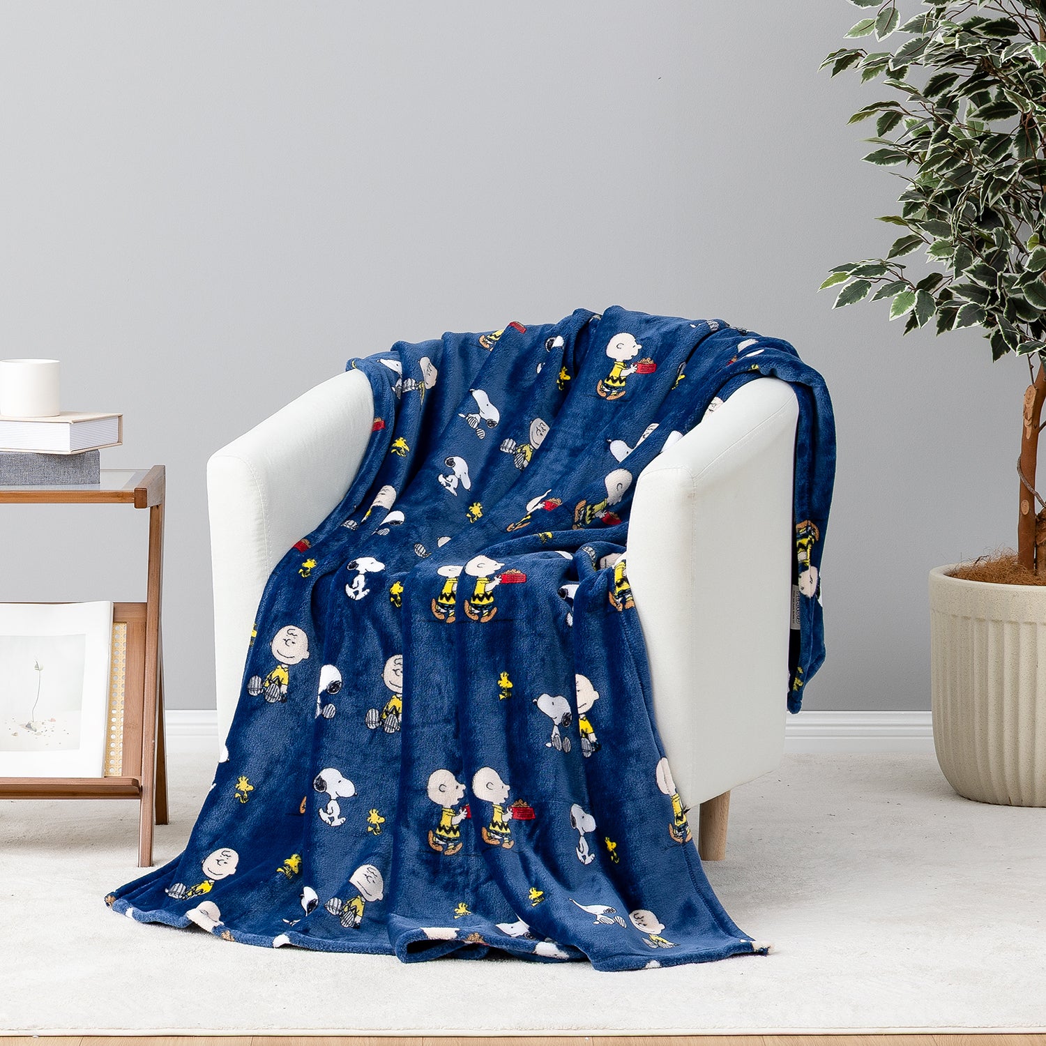 A navy blue plush throw blanket with a repeating cartoon character print is draped over a white armchair in a styled room setting.