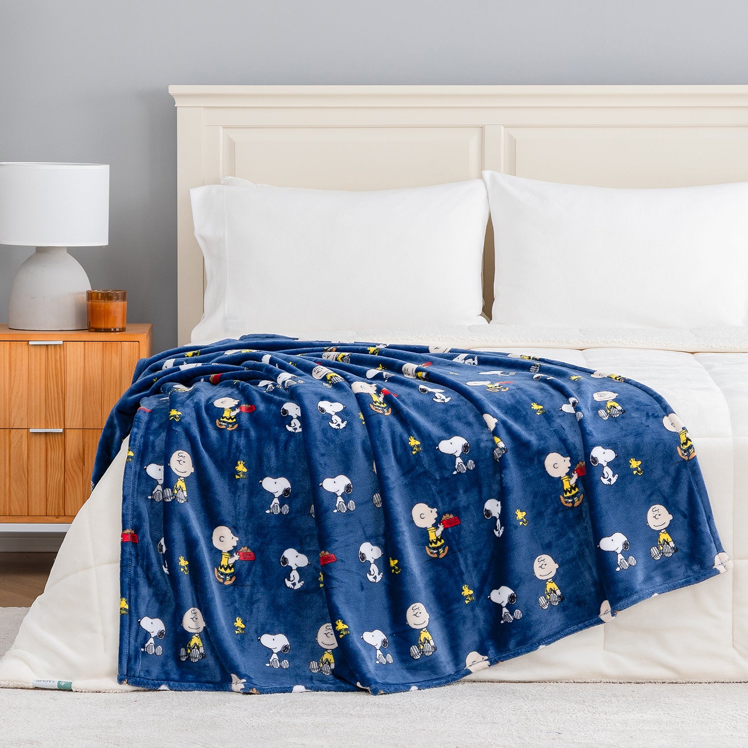 A navy blue fleece throw blanket featuring a repeating cartoon print of Snoopy, Woodstock, and other Peanuts characters, draped over a bed with white bedding in a bedroom setting.