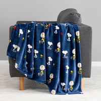A navy blue plush throw blanket featuring a repeating cartoon print of Snoopy, Woodstock, and Peanuts characters, draped over a gray armchair in a clean indoor setting.