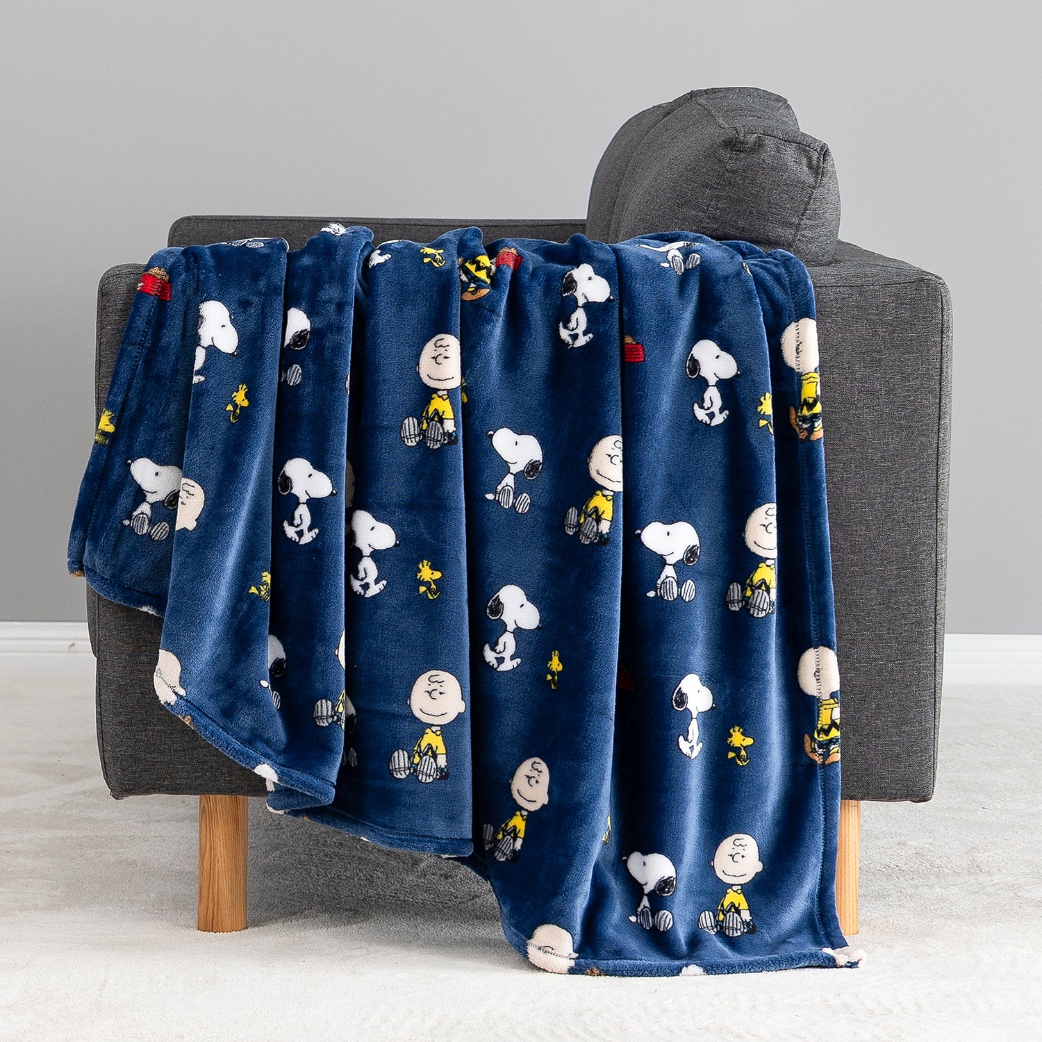 A navy blue plush throw blanket featuring a repeating cartoon print of Snoopy, Woodstock, and Peanuts characters, draped over a gray armchair in a clean indoor setting.