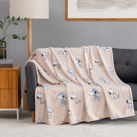 A light blush pink fleece throw blanket with an all-over cartoon Snoopy print, draped over a dark gray sofa in a modern living room setting.