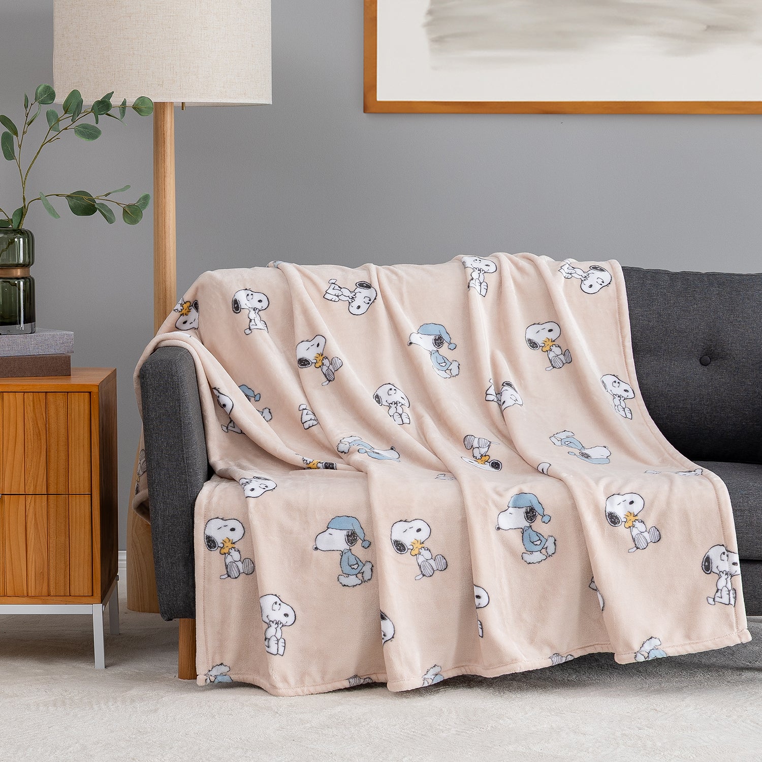 A light blush pink fleece throw blanket with an all-over cartoon Snoopy print, draped over a dark gray sofa in a modern living room setting.