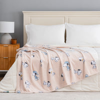 A light pink blanket with a repeating cartoon dog print is draped over a bed with white bedding and pillows in a bedroom setting.