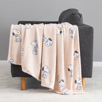 A light peach fleece throw blanket with a repeating Snoopy cartoon print is draped over a dark gray armchair with wooden legs in a clean studio setting.