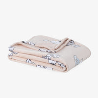 A folded plush blanket in a light peach-beige color, featuring a cartoon dog pattern in white and light blue, set against a plain white background.
