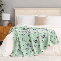 Bed with a green blanket featuring cartoon characters, white pillows, and a wooden nightstand with a lamp.