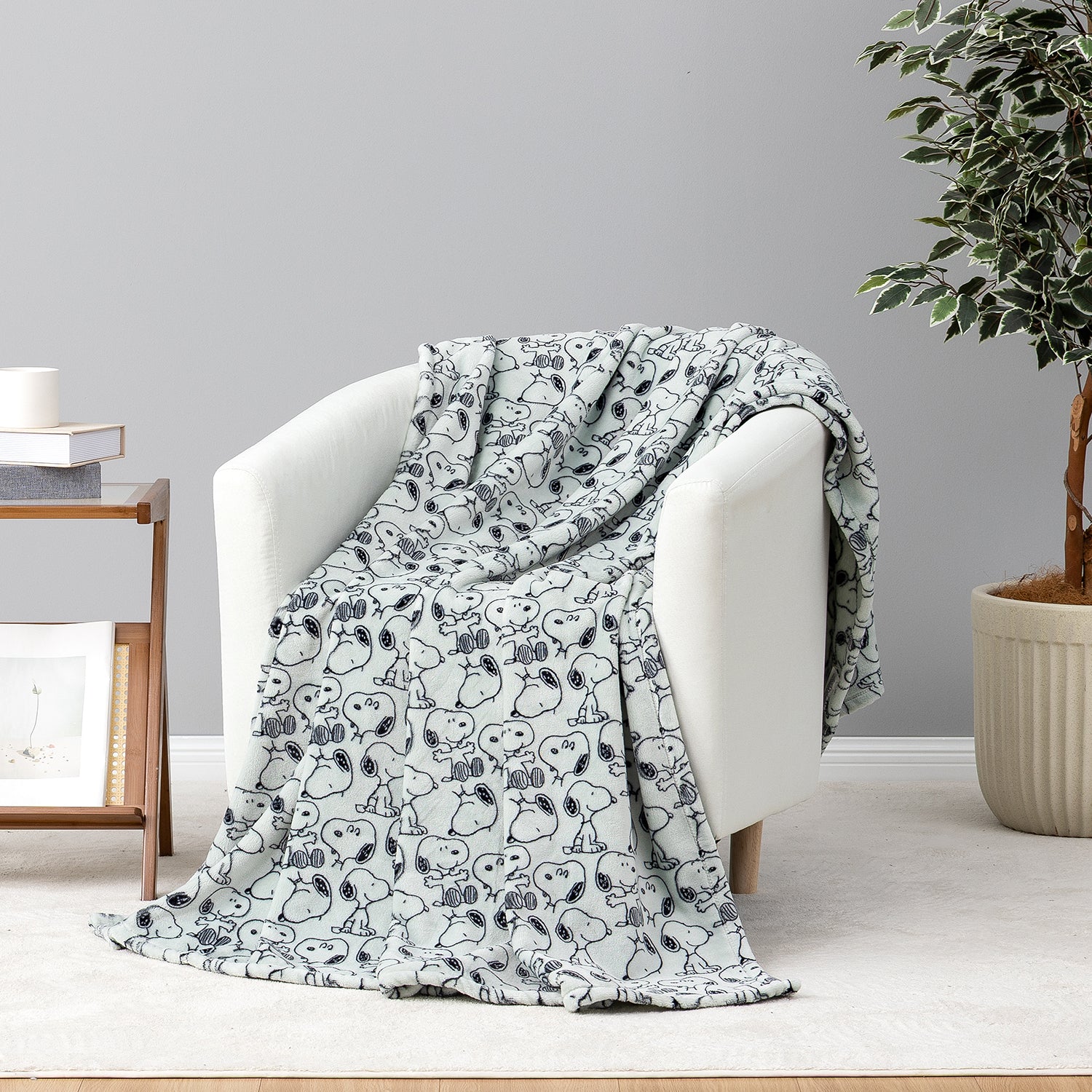 A light gray throw blanket with a black Snoopy and Woodstock cartoon pattern draped over a white armchair in a styled room setting.