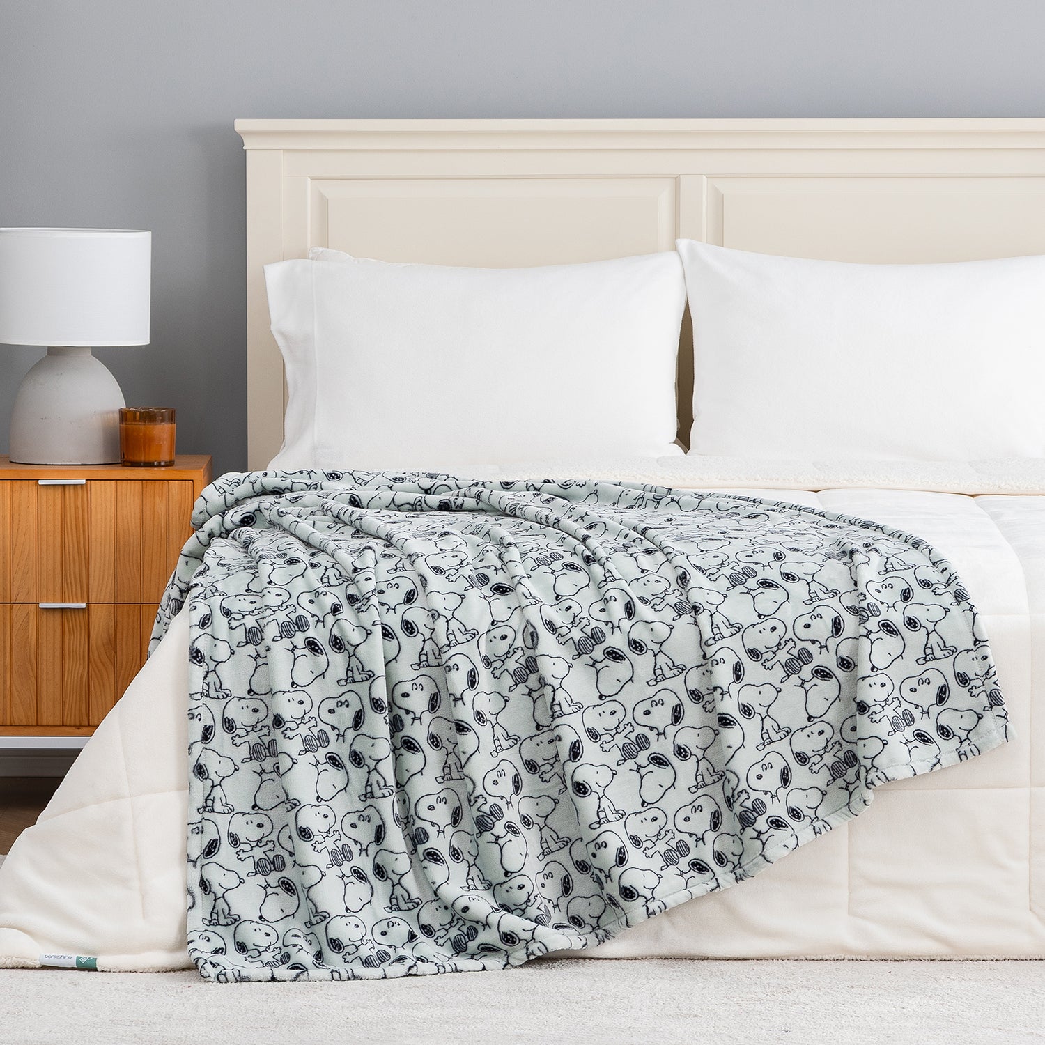 A light gray blanket with a black-and-white cartoon dog pattern is draped over a bed with white bedding and pillows in a bedroom setting.