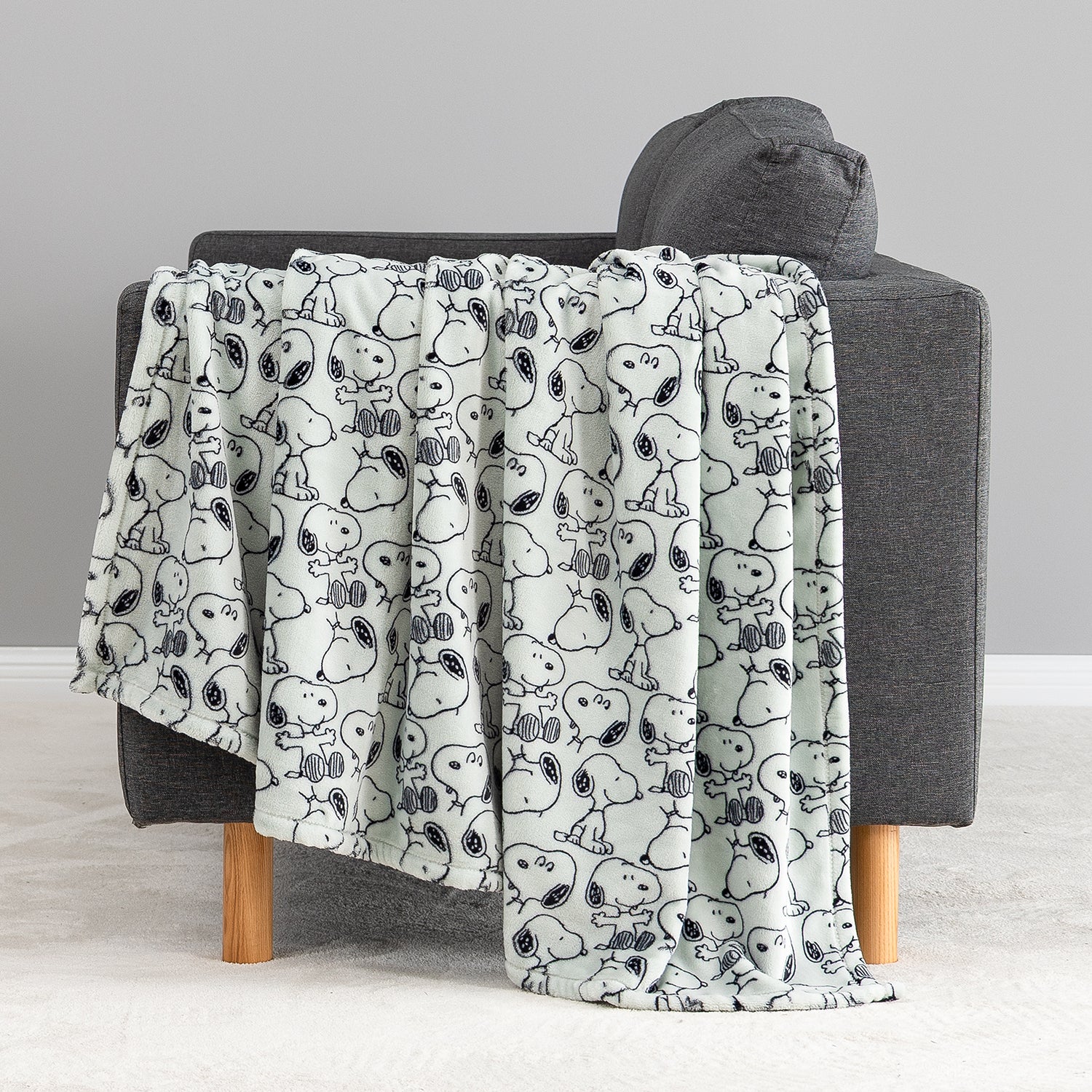 A light gray throw blanket with a black-and-white cartoon dog pattern is draped over a dark gray armchair with light wood legs in a minimalist room setting.