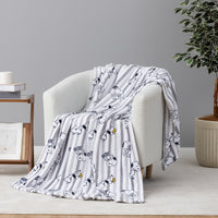 A white throw blanket with gray vertical stripes and cartoon dog illustrations is draped over a light gray armchair in a styled room setting with a side table and potted plant.