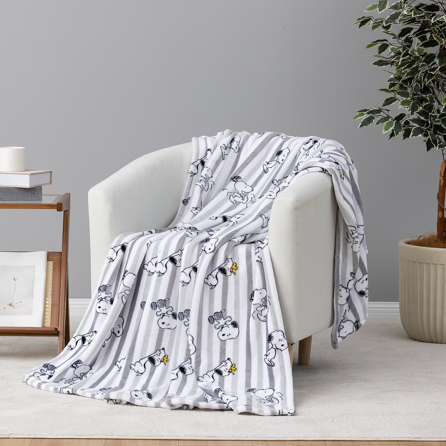 A white throw blanket with gray vertical stripes and cartoon dog illustrations is draped over a light gray armchair in a styled room setting with a side table and potted plant.