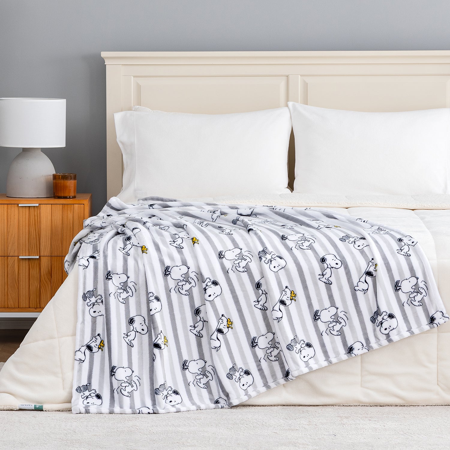 A striped throw blanket with a cartoon dog print draped over a bed in a styled bedroom setting. The blanket features white and gray vertical stripes with black-and-white cartoon characters and small yellow accents, set against a neutral gray wall and beige bed frame.