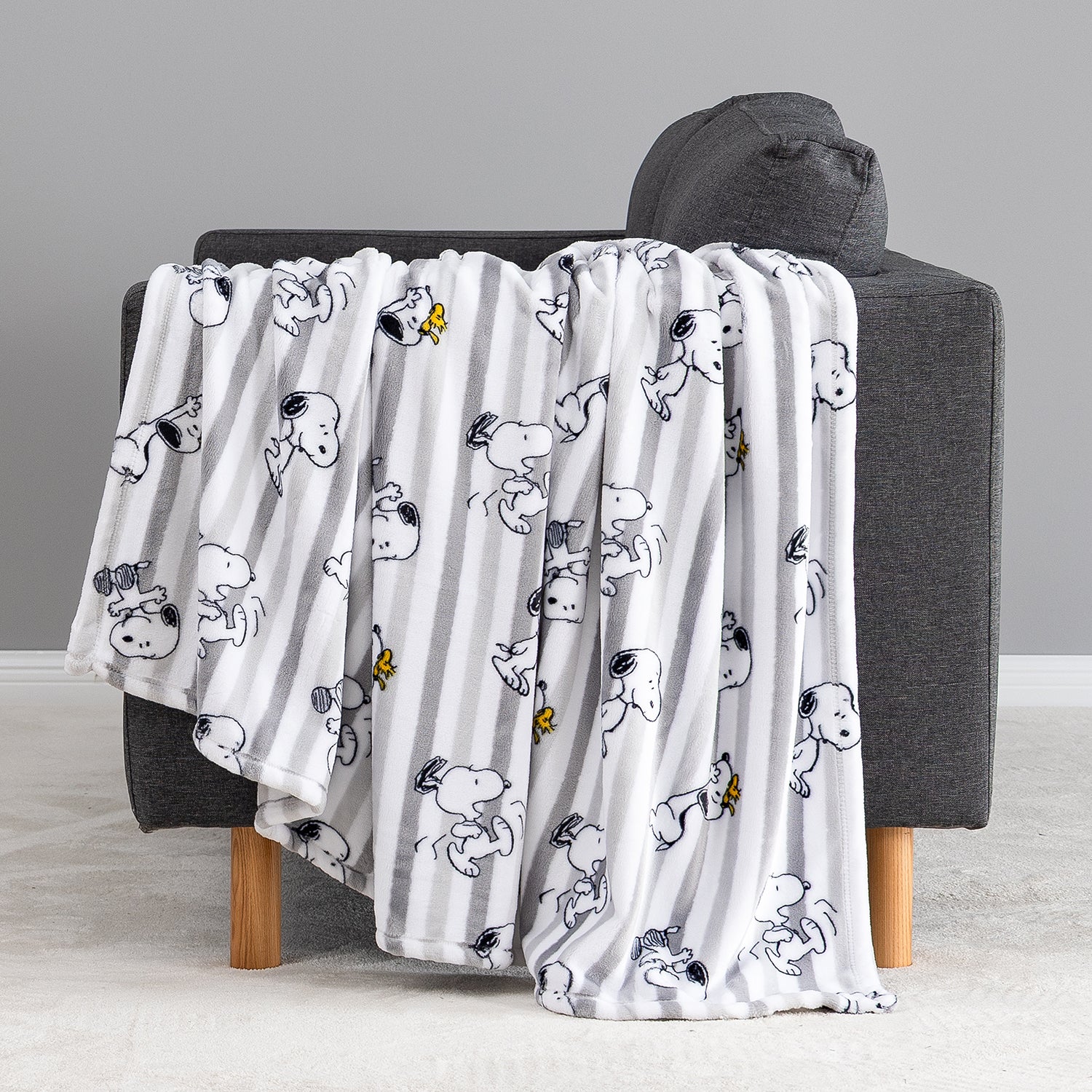 A white throw blanket with vertical gray stripes and a repeating cartoon dog print, draped over a dark gray armchair with wooden legs in a clean indoor setting.