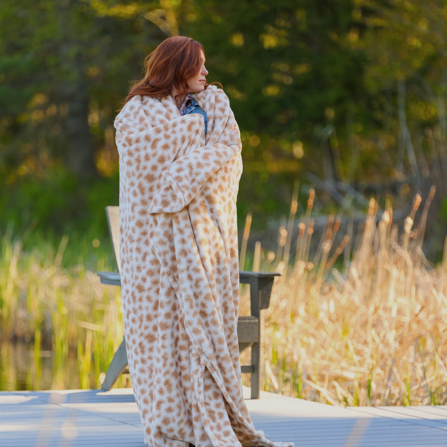 Person wrapped in a patterned blanket standing outdoors near water and tall grass.