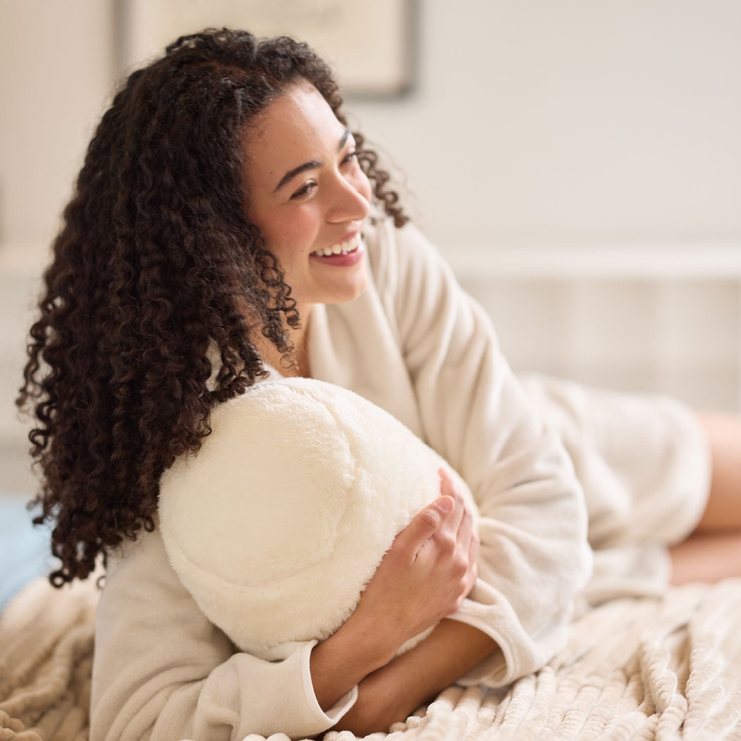 Woman in a robe holding a fluffy white pillow in a cozy room.