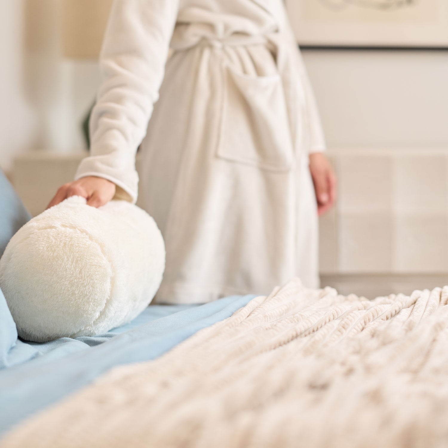 Person in a bathrobe holding a fluffy white pillow on a bed.