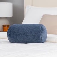 Blue cylindrical pillow on a bed with white bedding and a lamp in the background.