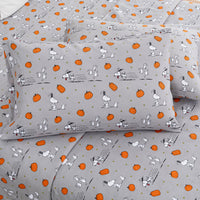 A gray bedding set with a repeating print of Snoopy and Woodstock alongside orange pumpkins and small gold dots, shown on a bed with two matching pillowcases.
