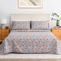 A bed dressed in a light gray patterned bedding set with matching pillowcases, featuring a repeating print of cartoon characters and orange circular motifs. The bed is styled in a bright bedroom with a cream headboard, wooden nightstands, a white lamp, and a green plant.