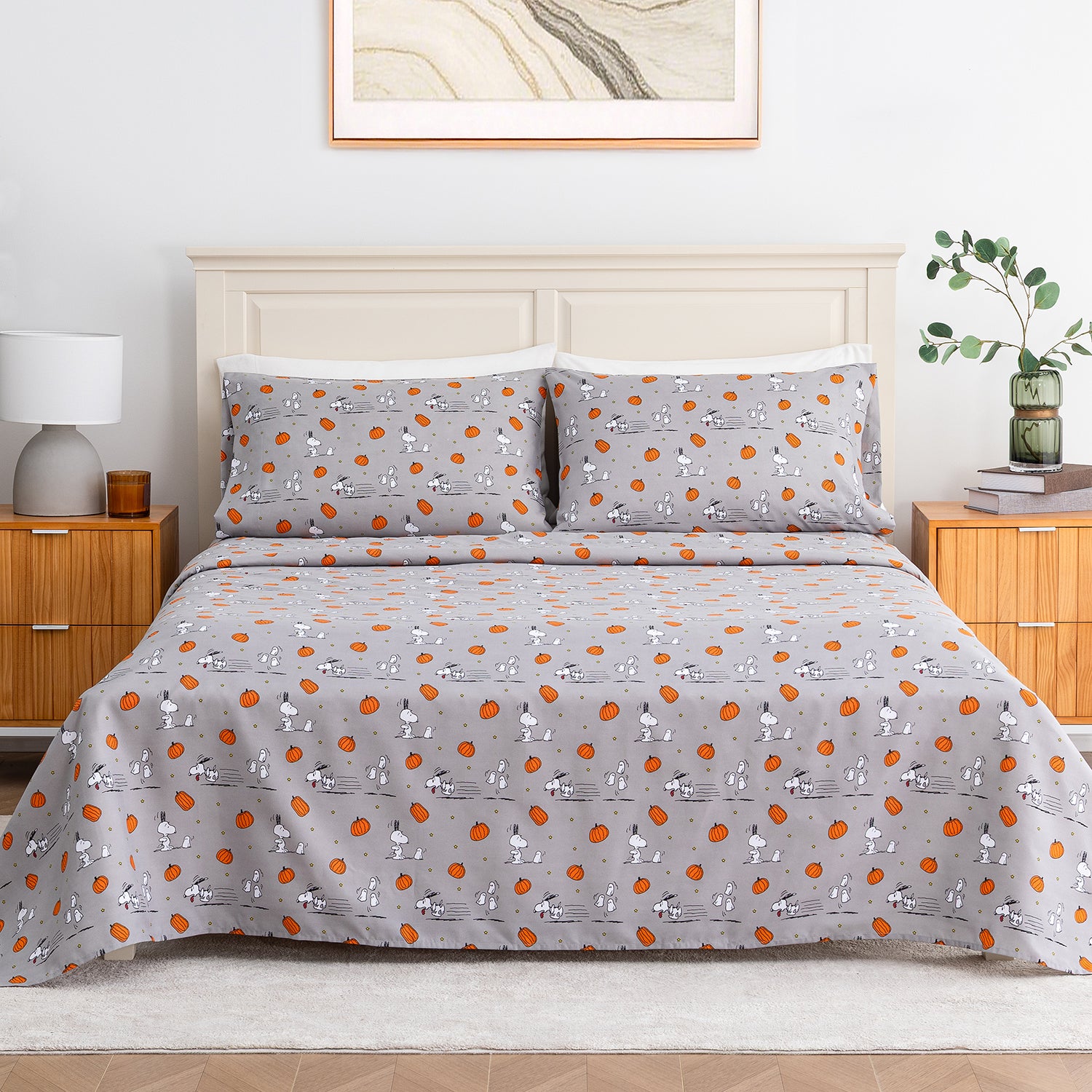 A bed dressed in a light gray patterned bedding set with matching pillowcases, featuring a repeating print of cartoon characters and orange circular motifs. The bed is styled in a bright bedroom with a cream headboard, wooden nightstands, a white lamp, and a green plant.