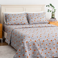 A bed dressed in a gray patterned bedding set featuring Snoopy and Woodstock illustrations with orange pumpkins, shown in a styled bedroom setting with a wooden headboard, bedside table, books, and a plant.