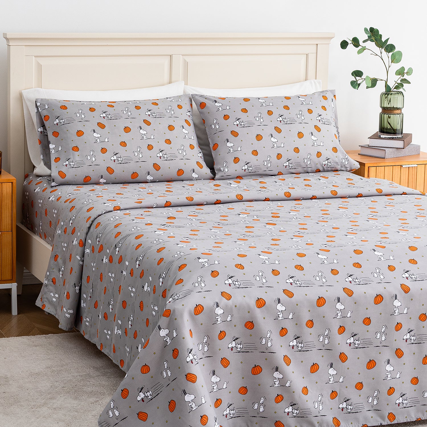 A bed dressed in a gray patterned bedding set featuring Snoopy and Woodstock illustrations with orange pumpkins, shown in a styled bedroom setting with a wooden headboard, bedside table, books, and a plant.