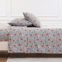 A gray bedding set featuring a bedspread and two matching pillowcases with a cartoon dog print in orange, white, and gray, styled on a bed against a white paneled wall.