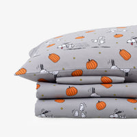 A stack of gray bedding, including a pillowcase and folded sheets, printed with an orange pumpkin and white cartoon dog pattern on a light gray background.