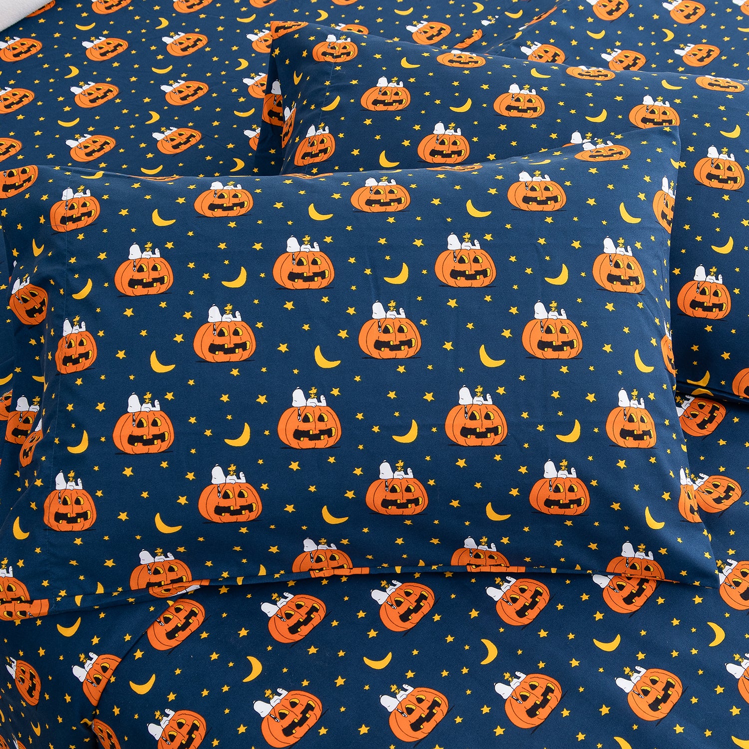 A close-up of a bedding set, likely pillowcases or a fitted sheet, featuring a Halloween pattern with orange jack-o'-lanterns, yellow crescent moons, and small yellow stars on a dark navy blue background.
