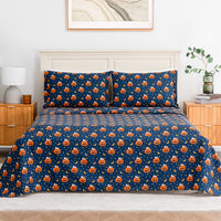 A neatly made bed in a bedroom setting featuring a navy blue bedding set with an all-over repeating pattern of orange jack-o'-lantern pumpkins and small white stars. The bed is styled with a cream headboard, wooden bedside tables, a lamp, and a small plant.