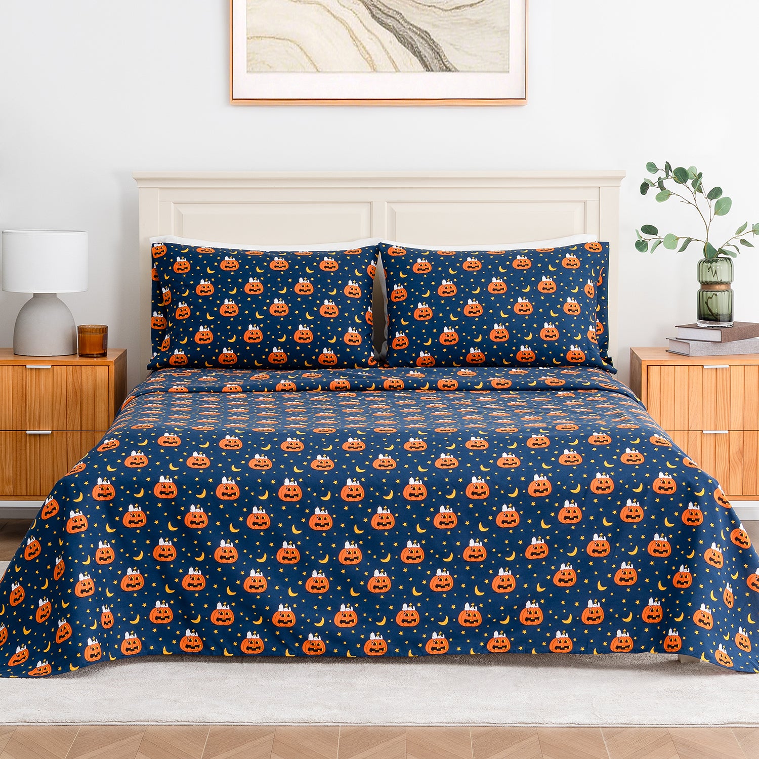 A neatly made bed in a bedroom setting featuring a navy blue bedding set with an all-over repeating pattern of orange jack-o'-lantern pumpkins and small white stars. The bed is styled with a cream headboard, wooden bedside tables, a lamp, and a small plant.