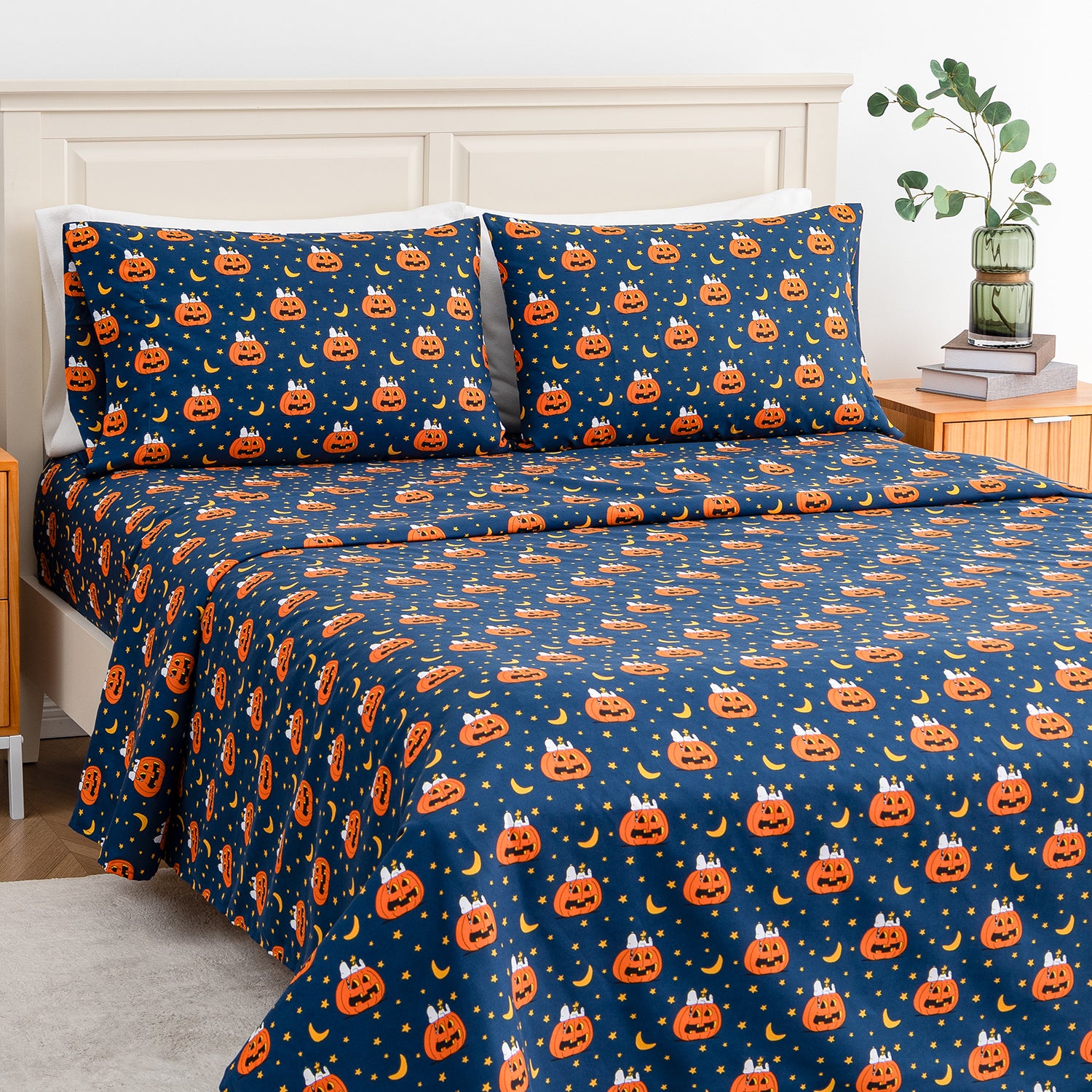 A bed dressed in a navy blue Halloween-themed bedding set with an all-over print of orange jack-o'-lanterns, yellow stars, and small white bats. The scene is styled in a bedroom with a light wood headboard, a wooden nightstand, books, a vase with greenery, and a light rug.