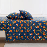 A bed dressed with a dark blue Halloween-themed bedding set featuring an orange jack-o'-lantern pattern, yellow crescent moons, and white stars, styled with two matching pillows against a white paneled wall.