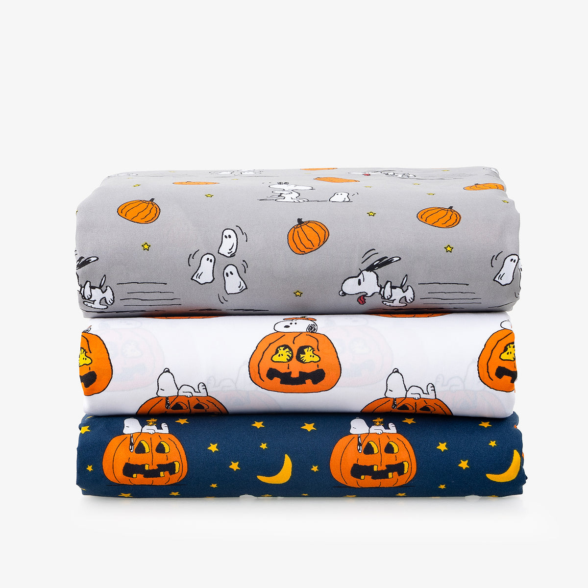 Three folded fabric items stacked vertically, each with a different Halloween-themed print: a gray Snoopy and Woodstock pattern with pumpkins and ghosts, a white Snoopy pattern with jack-o'-lanterns, and a navy blue Snoopy pattern with pumpkins, moons, and stars.
