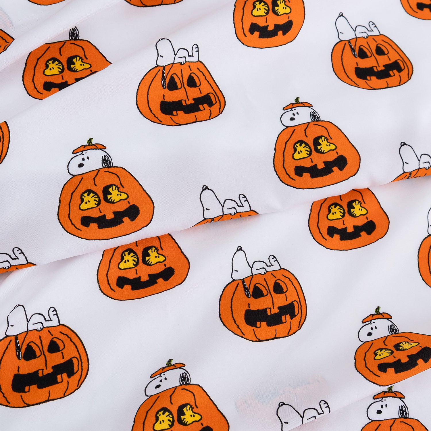 White fabric with a repeating pattern of orange jack-o'-lantern pumpkins and Snoopy lying on his back wearing a small orange hat, with Woodstock inside some pumpkins.