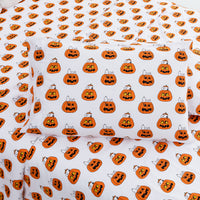 White bedding set with a repeating Halloween pumpkin print, including a pillowcase and sheets, featuring orange jack-o'-lanterns with black facial features and small white cats peeking out.