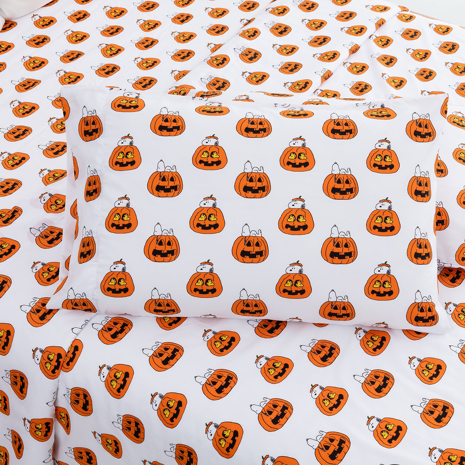 White bedding set with a repeating Halloween pumpkin print, including a pillowcase and sheets, featuring orange jack-o'-lanterns with black facial features and small white cats peeking out.