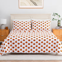 A bed dressed in a white bedding set with an all-over orange jack-o'-lantern pumpkin print, styled in a bright bedroom with wooden nightstands, a lamp, and a plant.