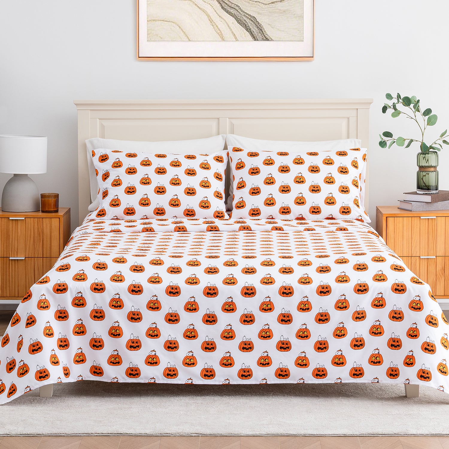 A bed dressed in a white bedding set with an all-over orange jack-o'-lantern pumpkin print, styled in a bright bedroom with wooden nightstands, a lamp, and a plant.