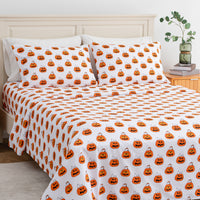 A bed dressed in a white bedding set with an all-over orange jack-o'-lantern pumpkin print, styled in a bedroom with a light wood headboard, bedside table, books, and a vase with greenery.