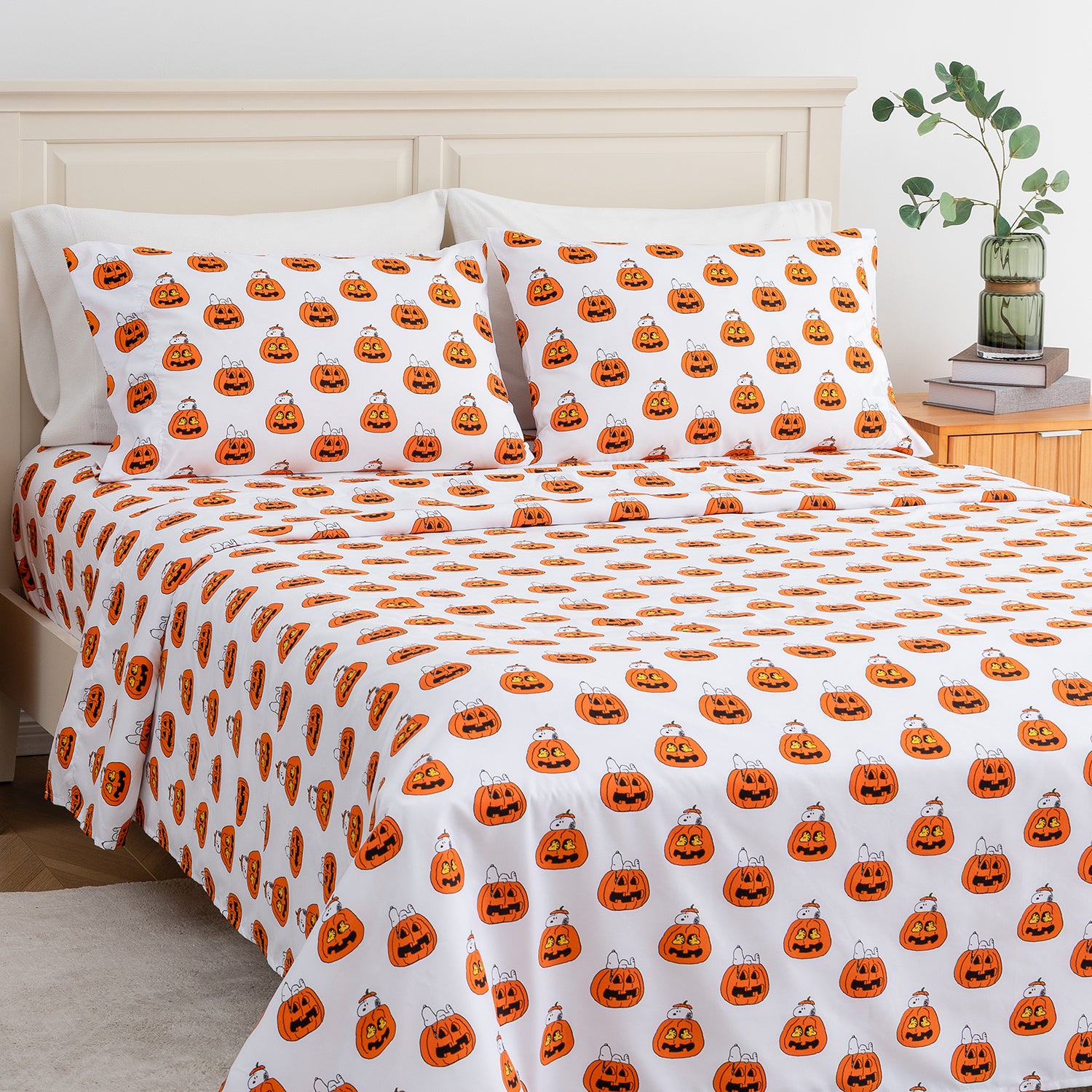 A bed dressed in a white bedding set with an all-over orange jack-o'-lantern pumpkin print, styled in a bedroom with a light wood headboard, bedside table, books, and a vase with greenery.