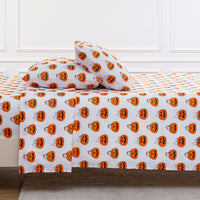 A bed dressed in a white sheet and pillowcase set printed with an all-over orange jack-o'-lantern pattern, shown in a styled bedroom setting with white paneled walls.