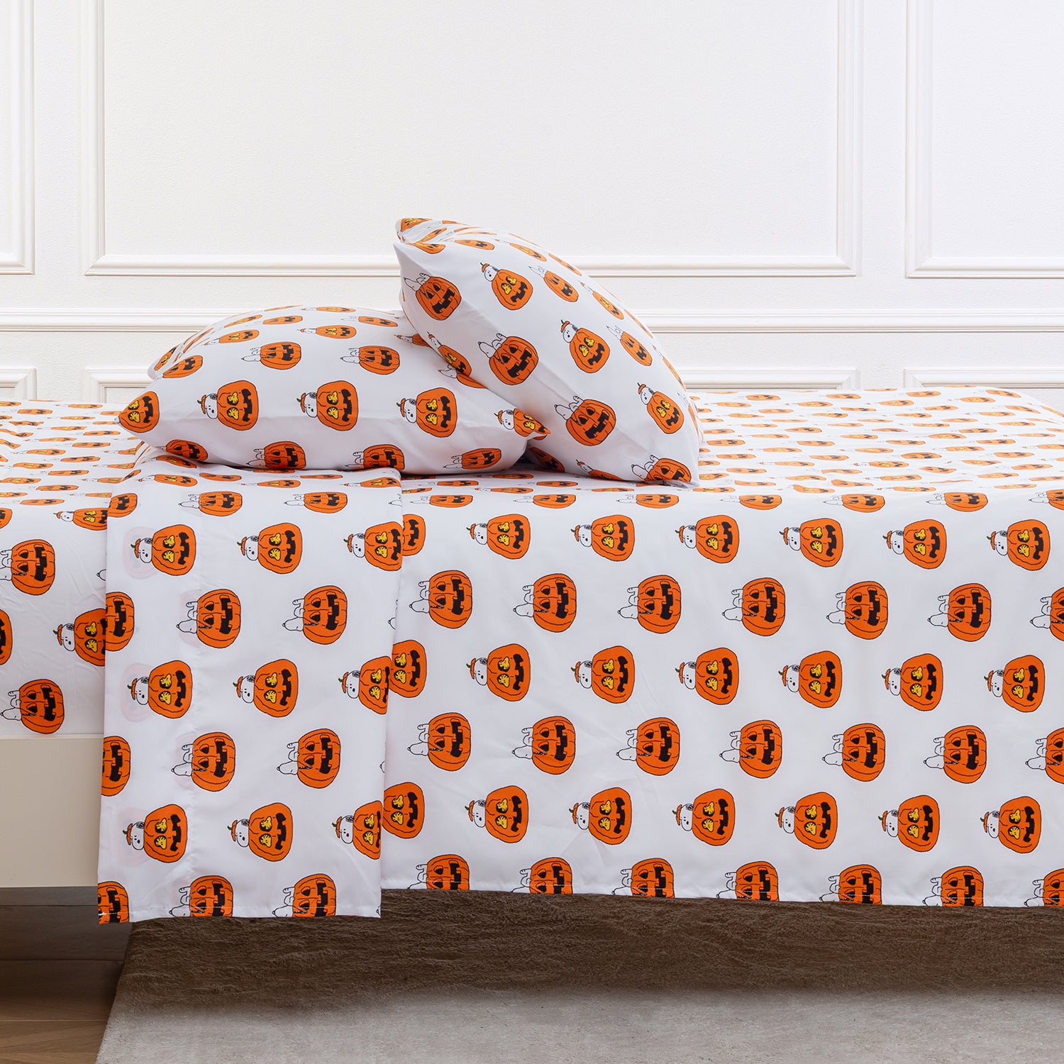 A bed dressed in a white sheet and pillowcase set printed with an all-over orange jack-o'-lantern pattern, shown in a styled bedroom setting with white paneled walls.