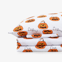 A folded white bedding set, likely a pillowcase and folded sheets, featuring a repeating Halloween pumpkin print with Snoopy and Wally from Peanuts.