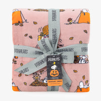 A folded pink Velvytoff throw blanket featuring a cartoon print of Snoopy and Woodstock with orange pumpkins and autumn leaves, wrapped in a gray ribbon printed with the 'PEANUTS' logo and a product tag.