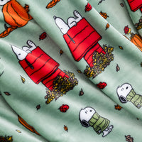 A close-up of a fabric surface featuring a repeating print of Snoopy and Woodstock in red and green sweaters, with pumpkins, leaves, and autumn foliage on a pale mint background.