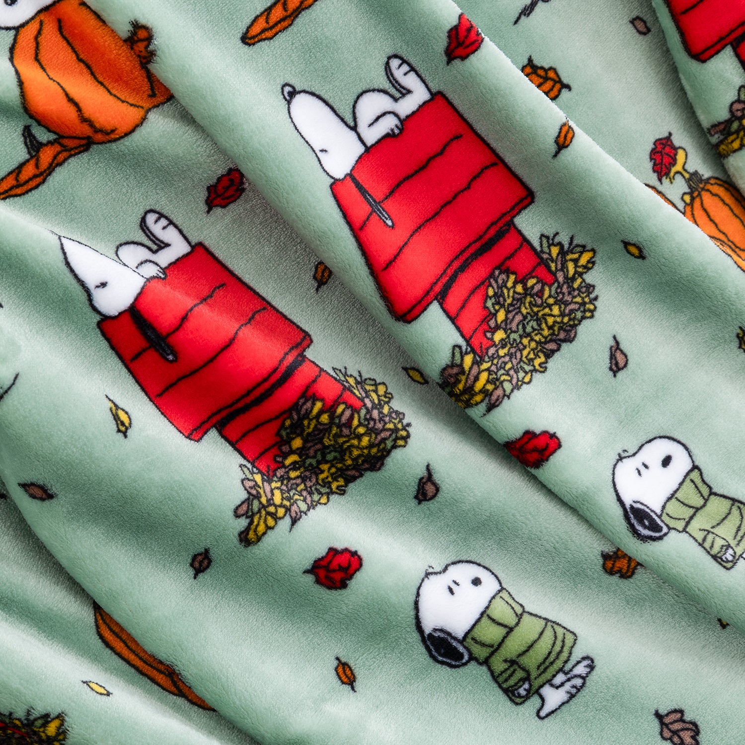 A close-up of a fabric surface featuring a repeating print of Snoopy and Woodstock in red and green sweaters, with pumpkins, leaves, and autumn foliage on a pale mint background.