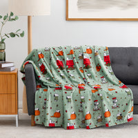 A sage green fleece throw blanket with a cartoon pumpkin and character print is draped over a gray sofa in a living room setting.